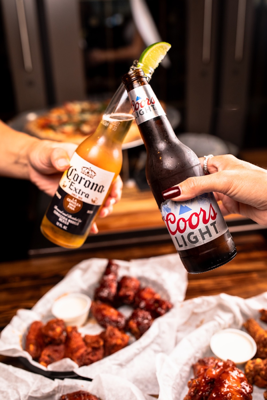 Two bottles of beer clinking over a basket of wings